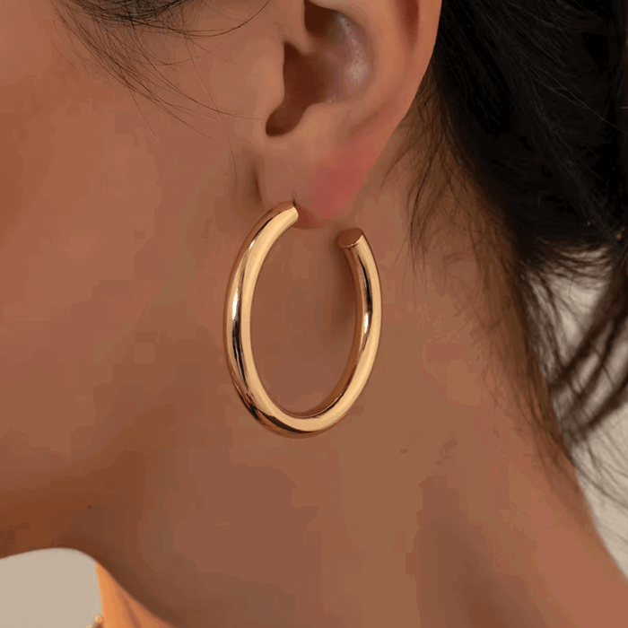 Gold-tone round hoop earrings worn on ear, thick profile with pin clasp visible.