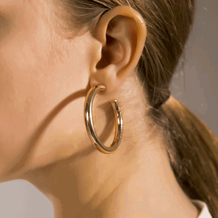 Gold-tone round hoop earrings worn on ear, larger thick hoops with pin clasp.