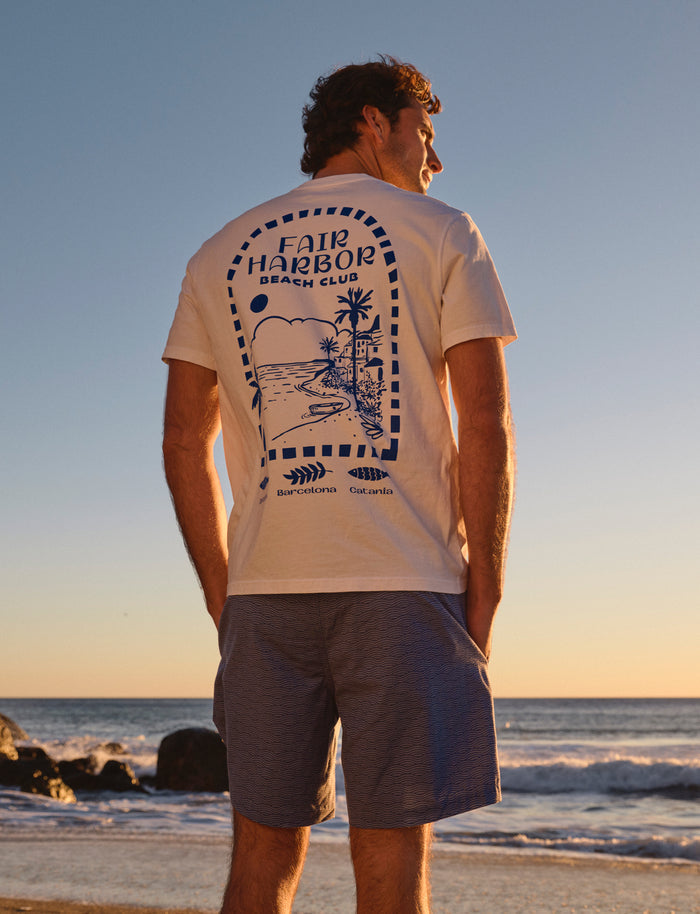 Person wearing a t-shirt with 'Fair Harbor Beach Club' design on a beach at sunset.