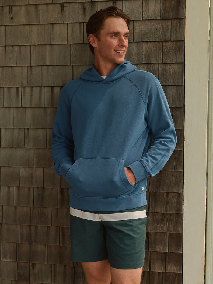 Man leaning on outside wall wearing blue hoodie with hands inside kangaroo pocket and green striped shorts