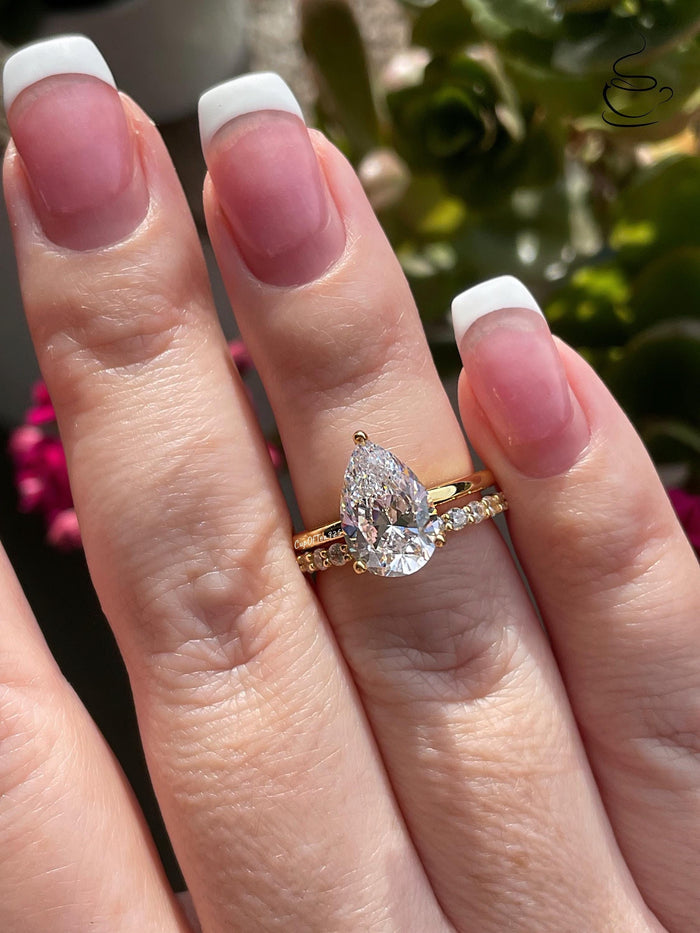 May include: A pear-shaped diamond engagement ring with a gold band and a smaller diamond band. The ring is set with a single large pear-shaped diamond in the center and a row of smaller diamonds on the band.